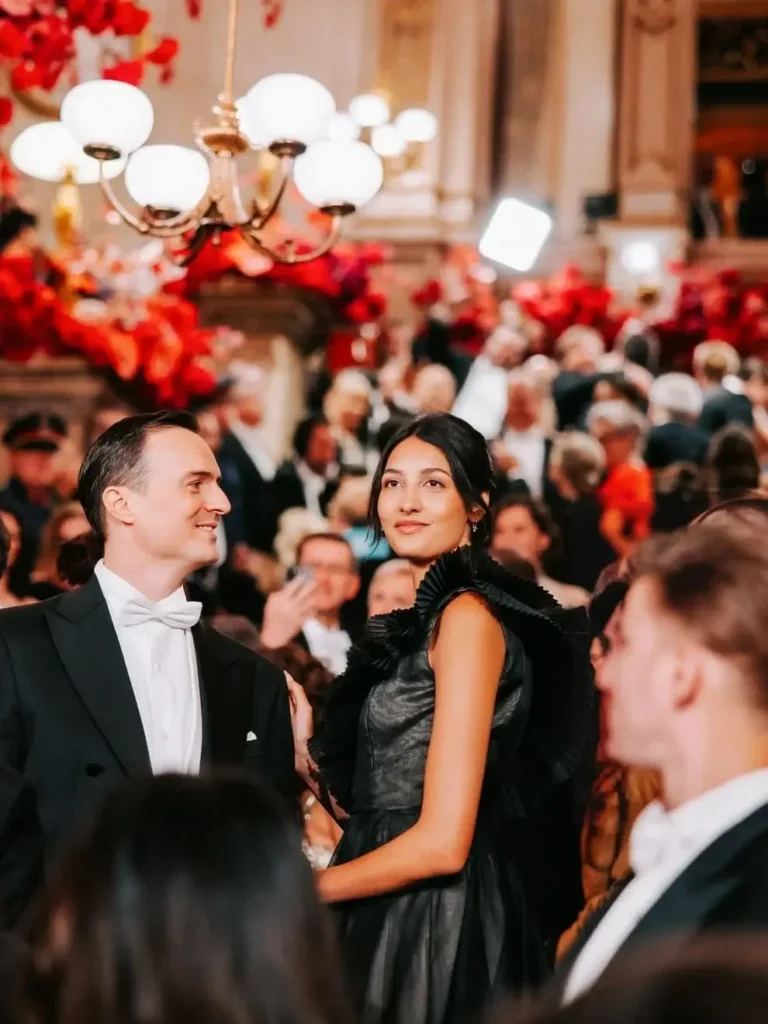 Ein gut gekleidetes Paar steht inmitten einer Menschenmenge auf einem glamourösen Nachtball, umgeben von Kronleuchtern und rotem Blumenschmuck im Hintergrund.